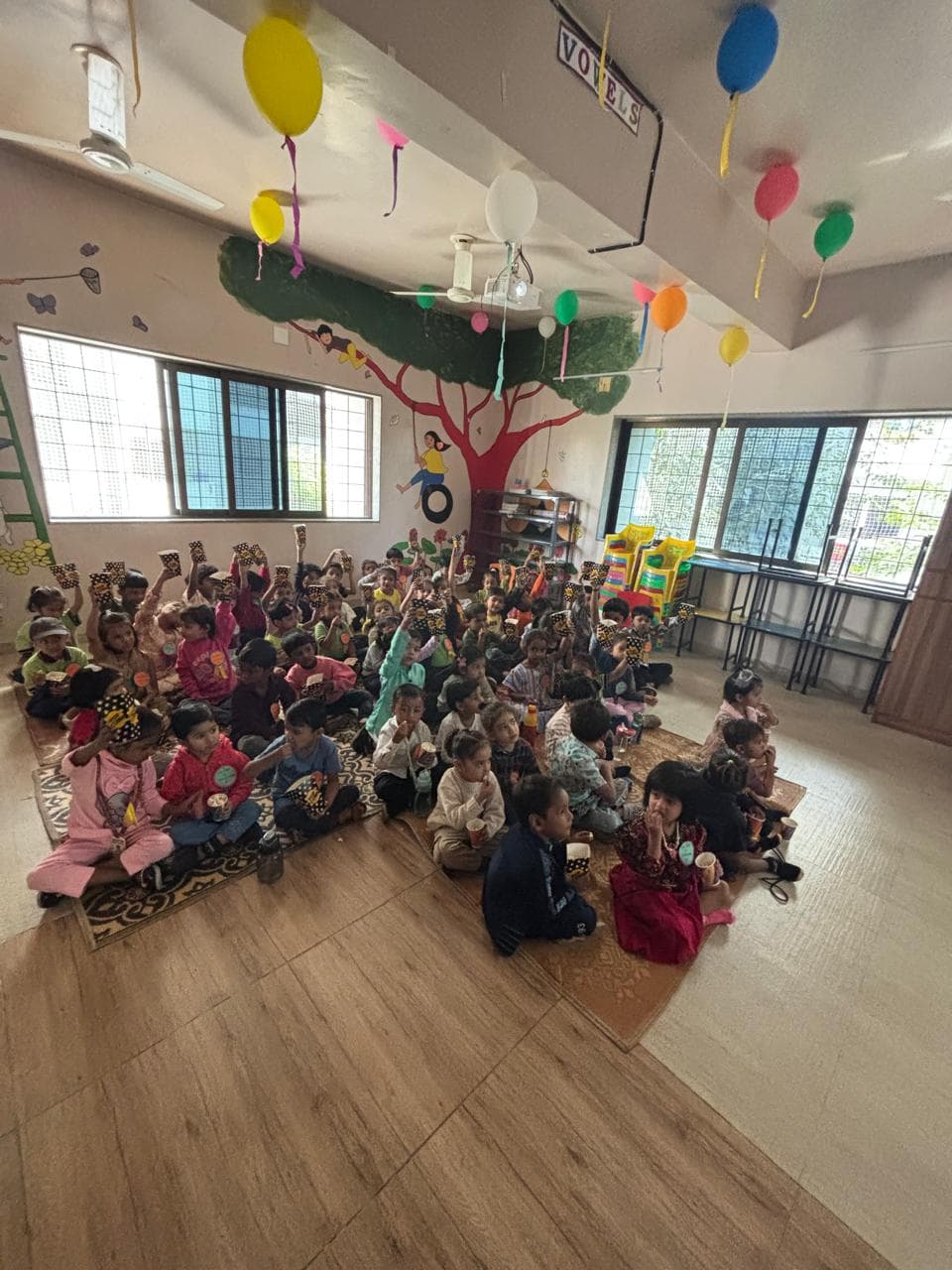 A large group of children seated together on mats in a spacious room decorated with balloons and a painted tree mural