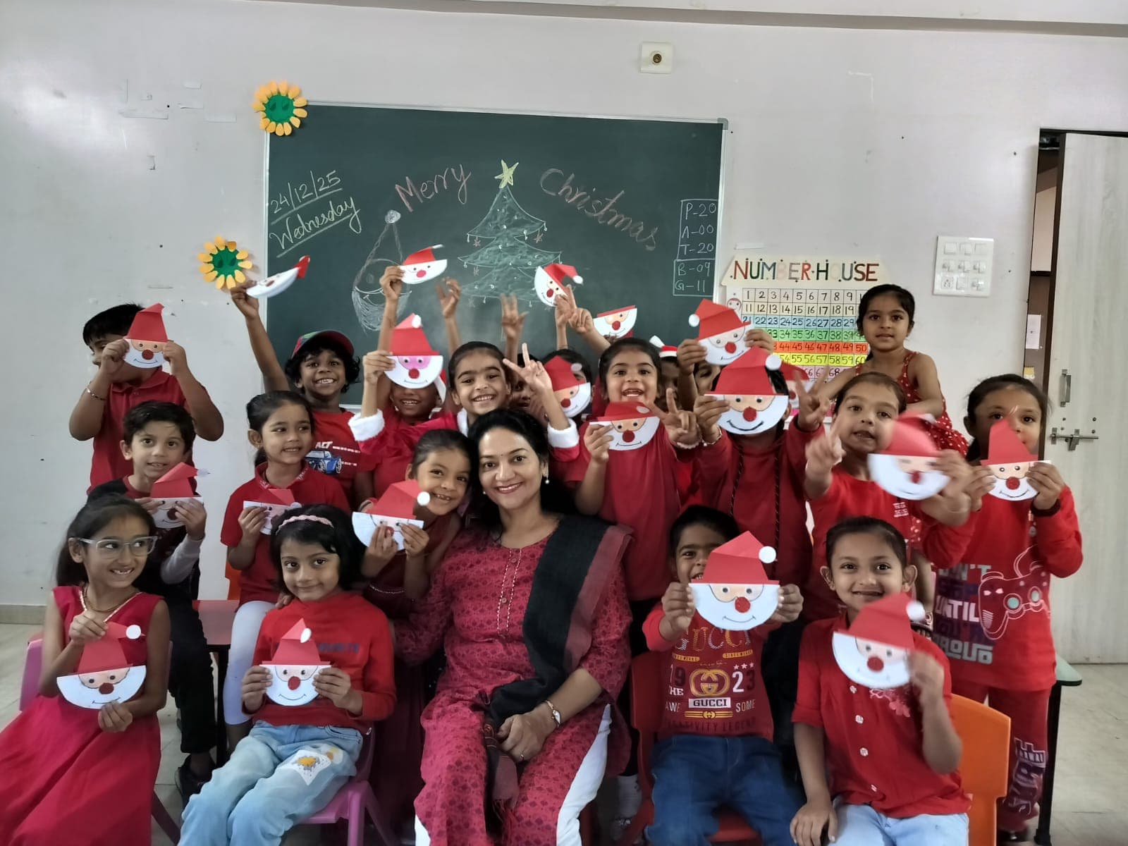 A smiling teacher with children dressed in red, holding up paper Santa crafts in front of a Merry Christmas chalkboard