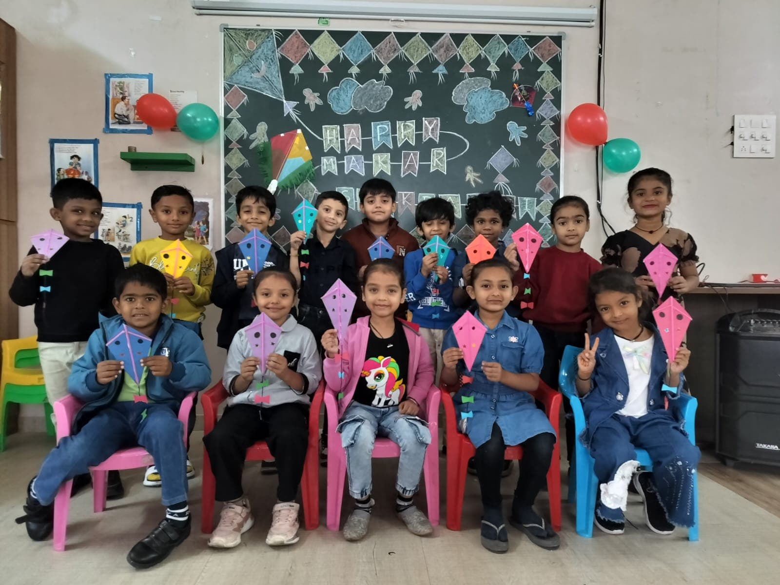Children holding colourful paper kites in front of a Happy Makar Sankranti chalkboard drawing
